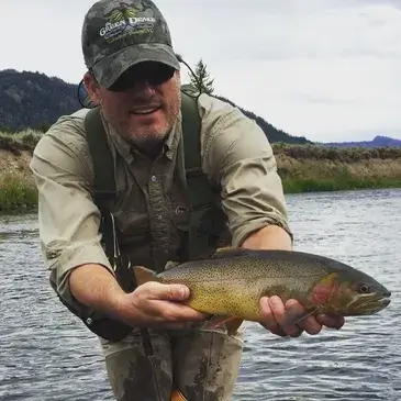 A Smokehouse Fly Fishing guide wearing a Green Drake hat smiling while holding a large trout, demonstrating the expert leadership provided on excursions to destinations like the Bahamas.