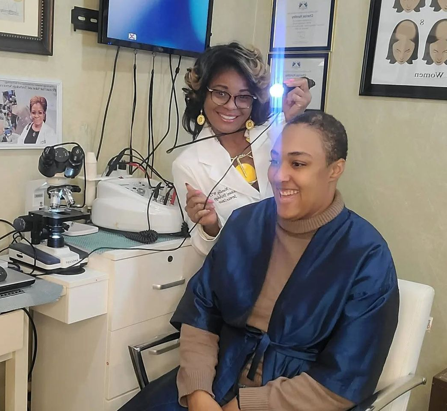 Trichology practitioner using a handheld diagnostic light to examine a seated client's scalp.
